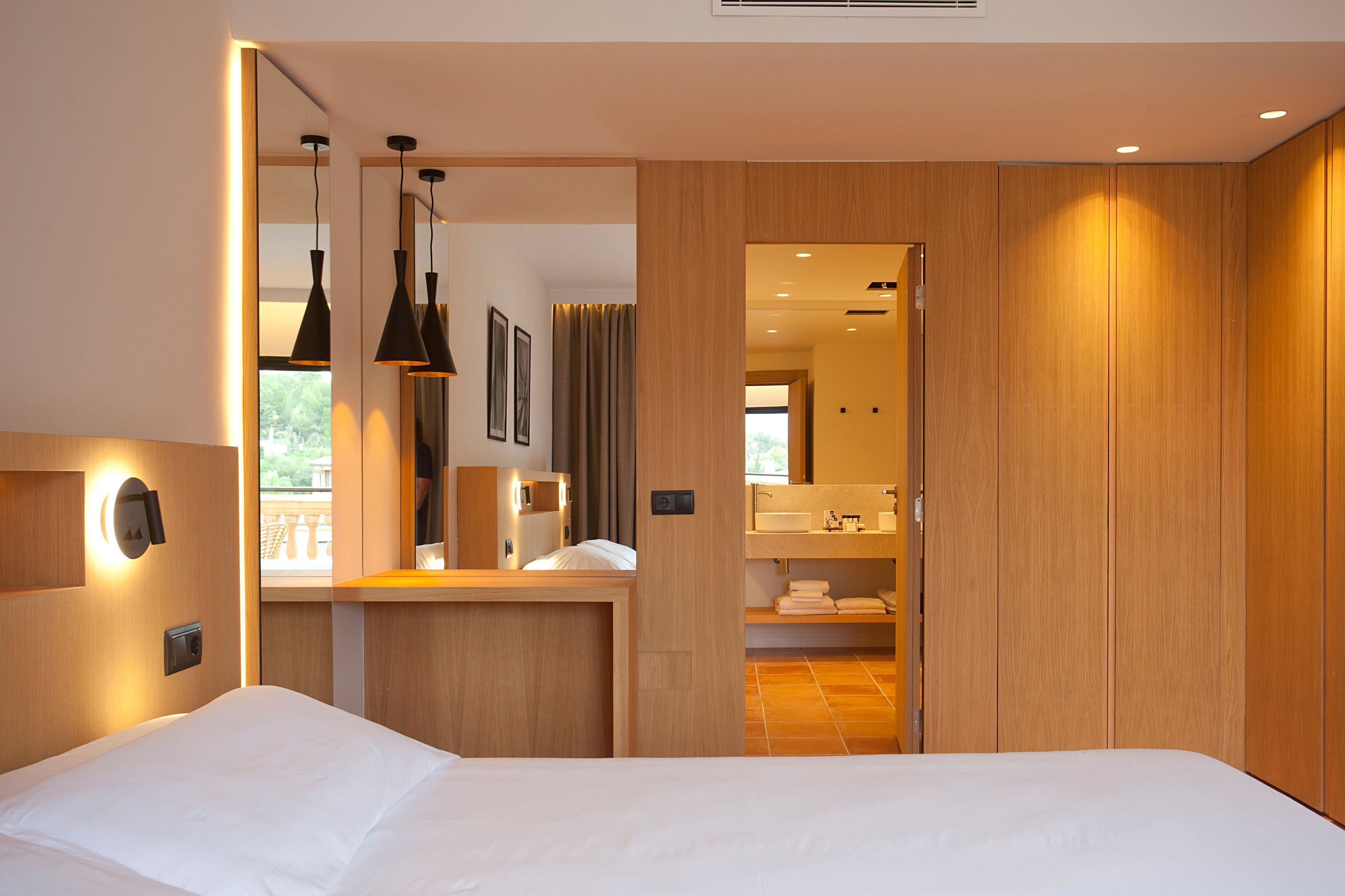 A modern hotel room with a bed, a nightstand, a mirror, and a bathroom visible through an open door.