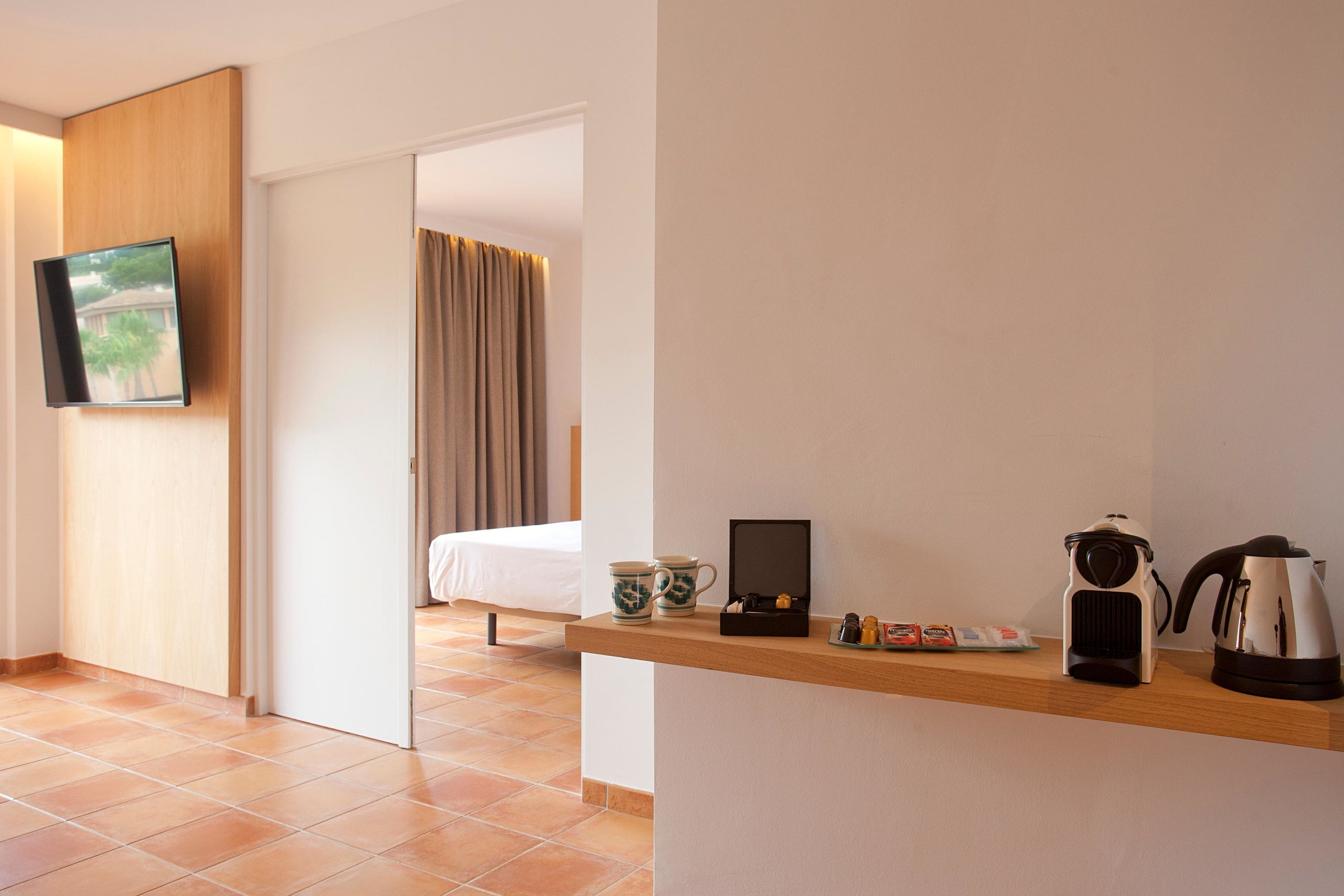 Hotelkamer met een bed, een houten plank met een waterkoker, kopjes en een klein doosje, en een aan de muur bevestigde televisie.