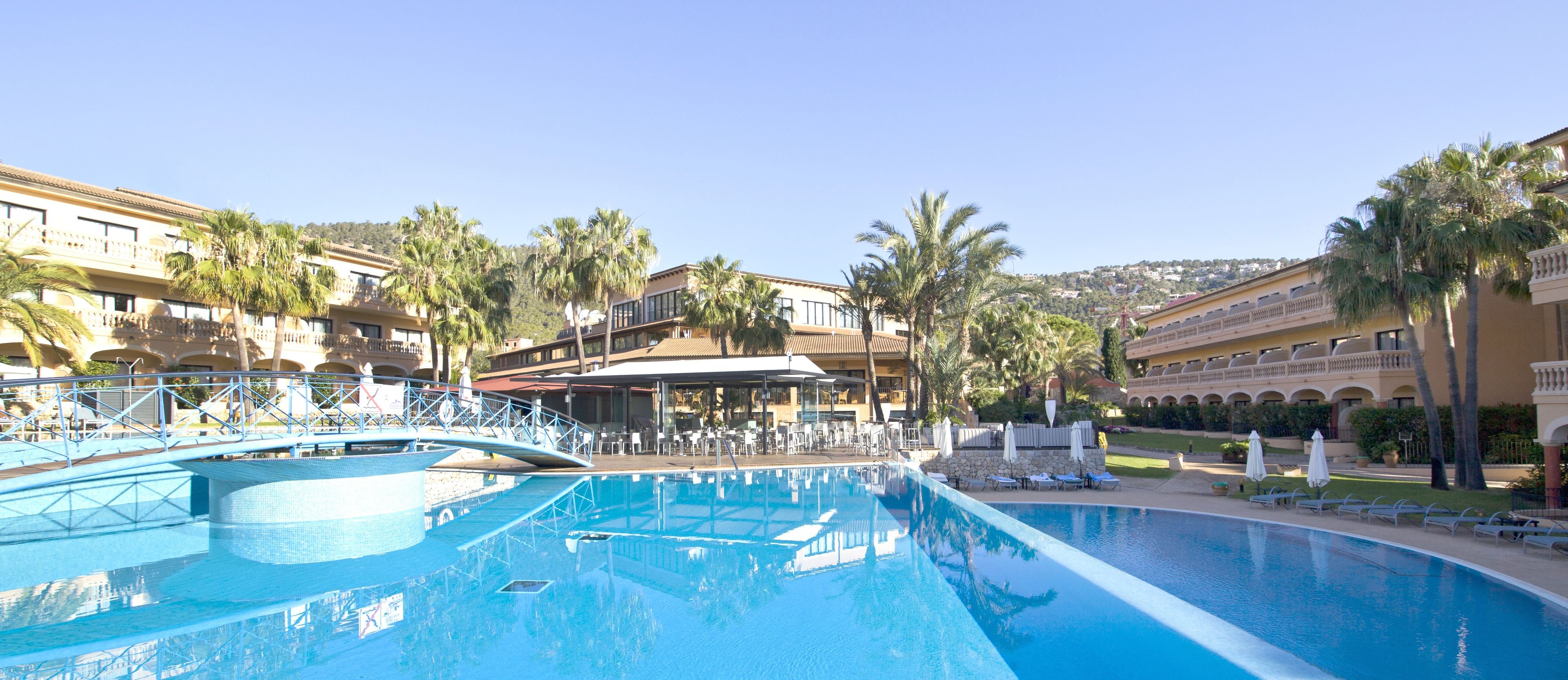 A large swimming pool with a bridge, palm trees, and a hotel with multiple balconies and outdoor seating areas.