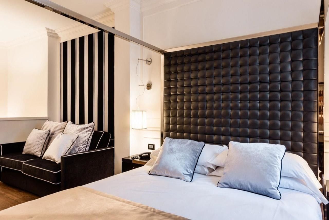 Ein modernes Hotelzimmer mit einem großen Bett, einem dunkel gepolsterten Kopfteil, einem Nachttisch mit Lampe sowie einer Sitzecke mit Sofa und Kissen.