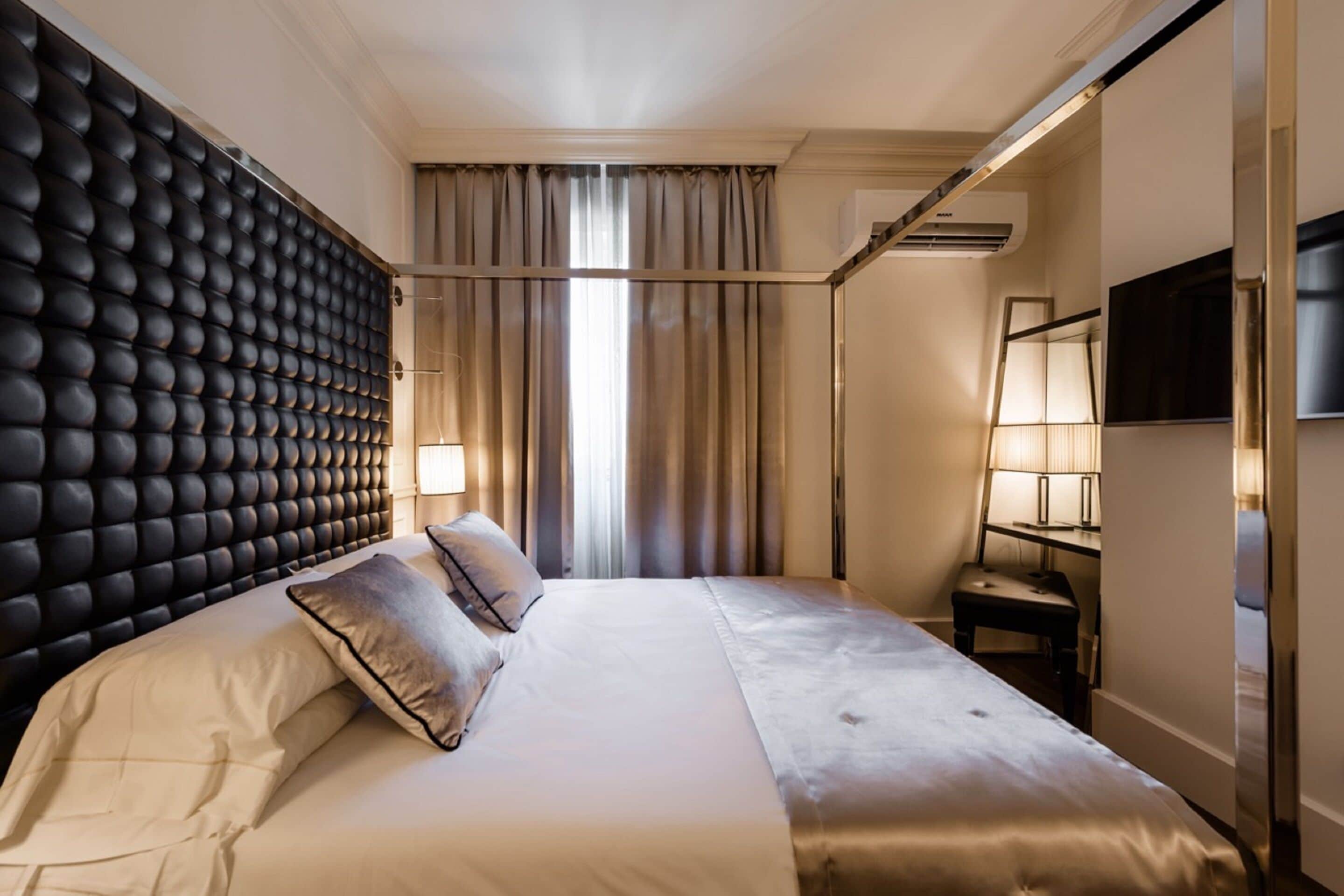 A hotel room with a tufted headboard, a large bed with white linens, a bedside table with a lamp, and a TV mounted on the wall.