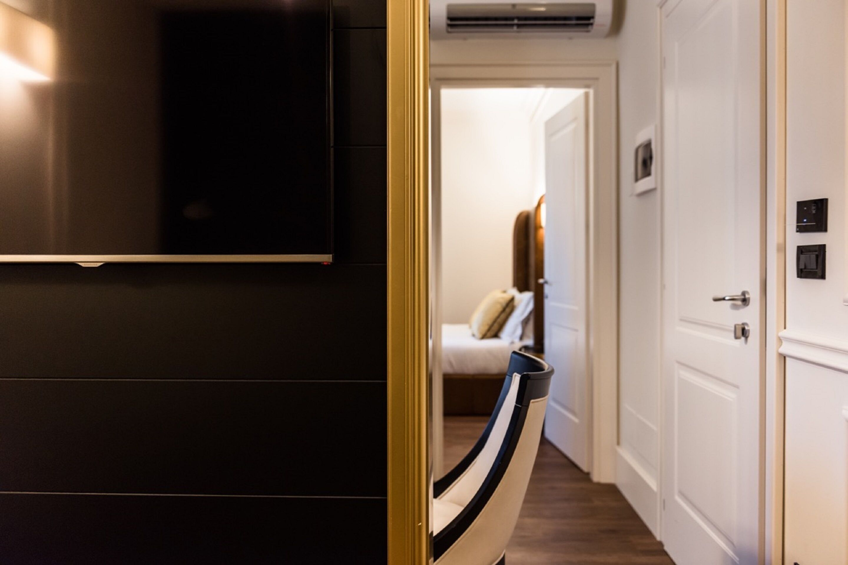 A hotel room with a dark-colored wardrobe, a television mounted on the wall, a bed with a white headboard, and a door leading to another room.