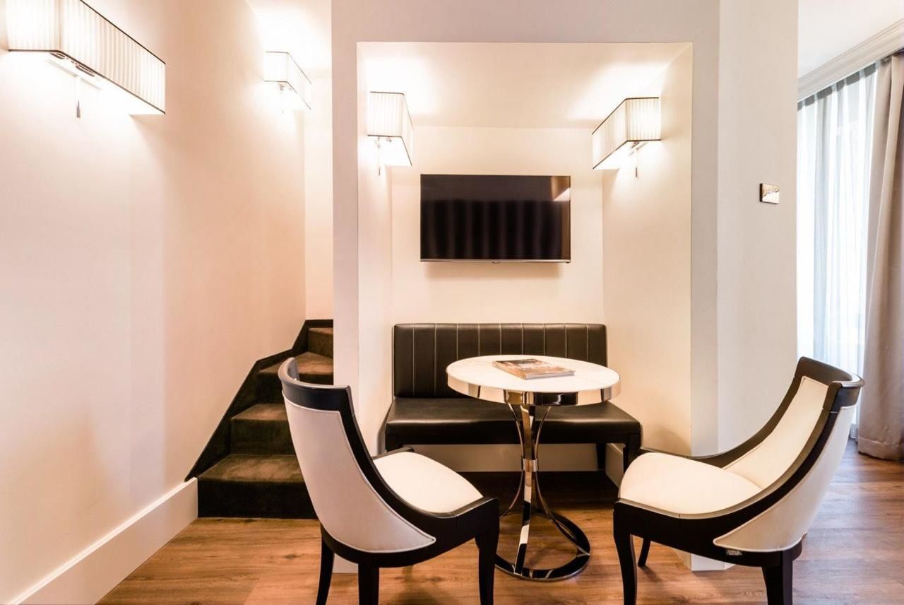 A modern hotel room with a black leather sofa, two white chairs, a small round table, and a flat-screen TV mounted on the wall.