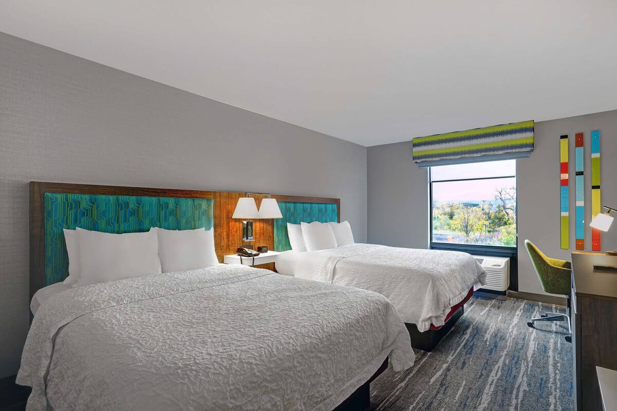 A hotel room with two beds, a desk, a chair, a window with a view, and a colorful wall decoration.
