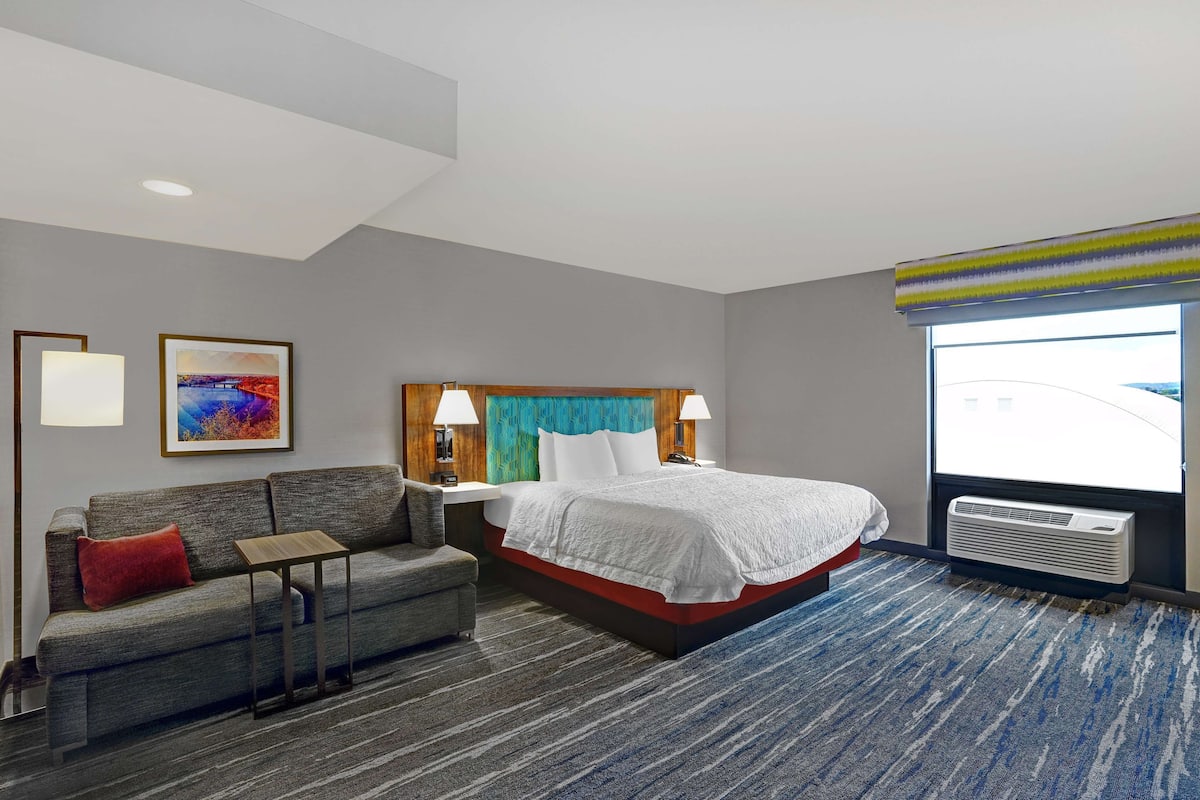 A hotel room with a bed, a sofa, a small table, a nightstand, a lamp, a window with a view, and a painting on the wall.