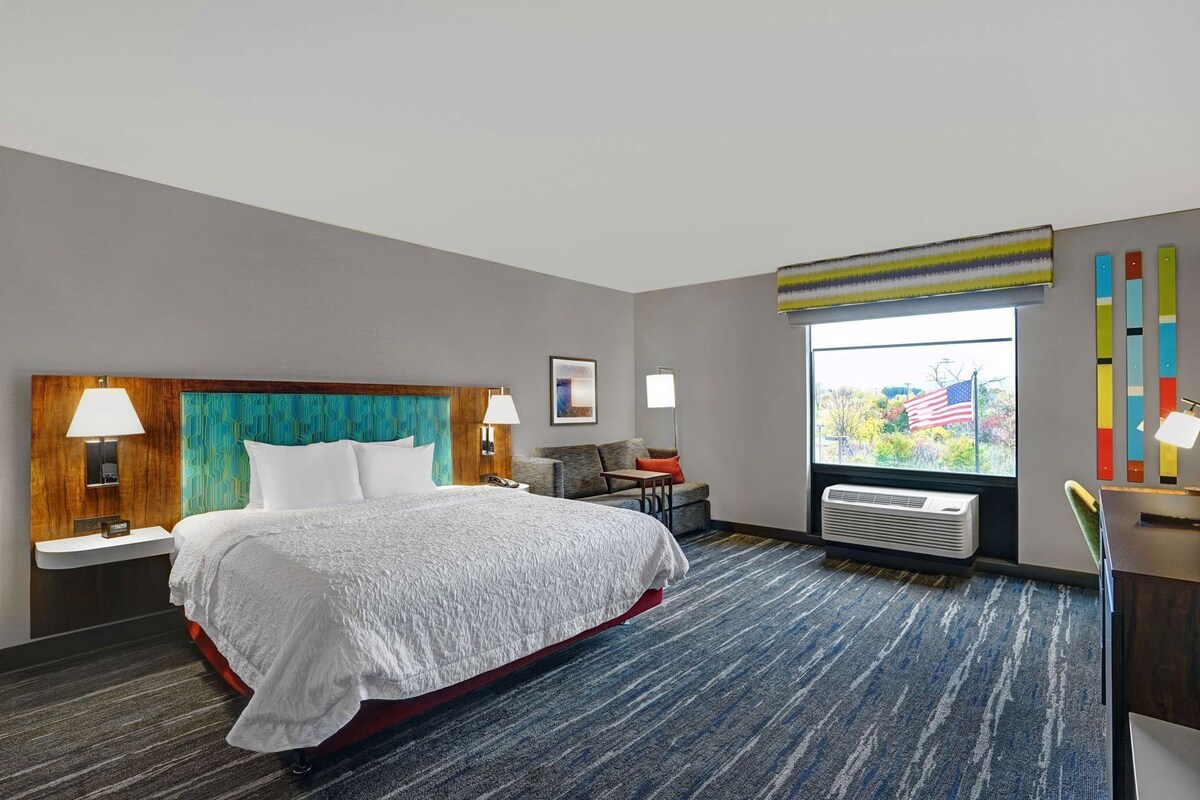 A hotel room with a bed, bedside tables, a desk, a chair, a window with a view, and a wall with colorful decor.
