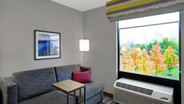 A modern living room with a grey sofa, a small wooden table, a red pillow, a floor lamp, and a large window with an air conditioning unit. - Hampton Inn Utica (Utica)