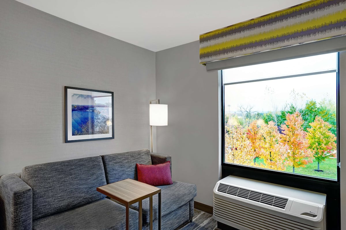A modern living room with a grey sofa, a small wooden table, a red pillow, a floor lamp, and a large window with an air conditioning unit.