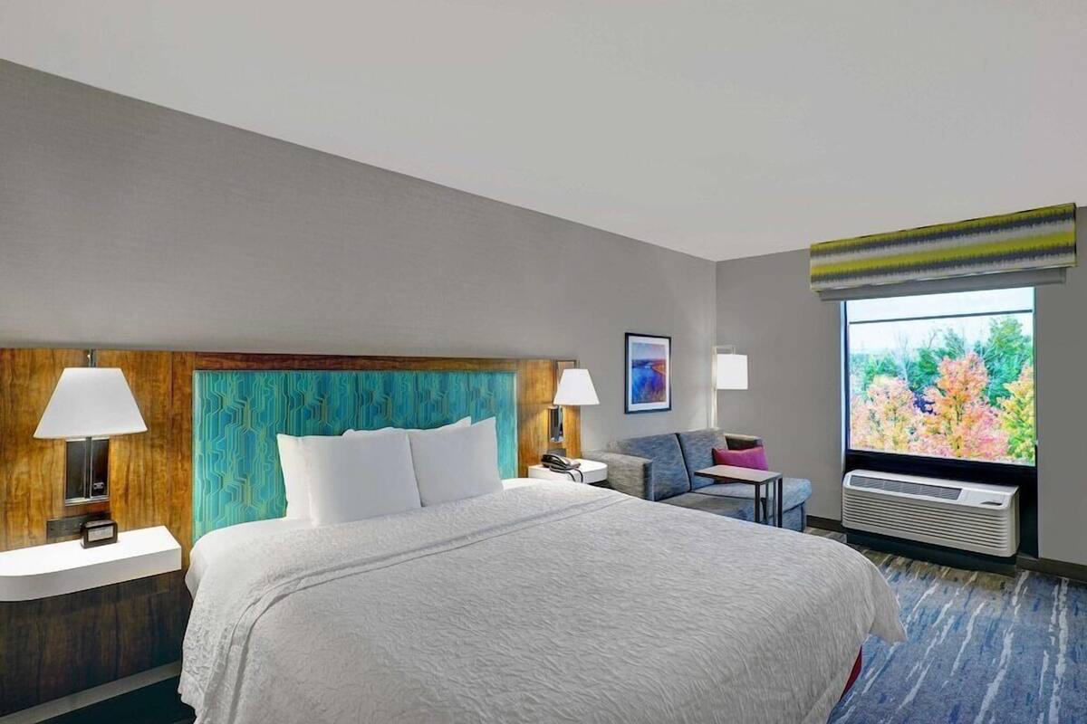 A hotel room with a large bed, two bedside tables with lamps, a small desk, a chair, a window with a view of greenery, and an air conditioning unit.
