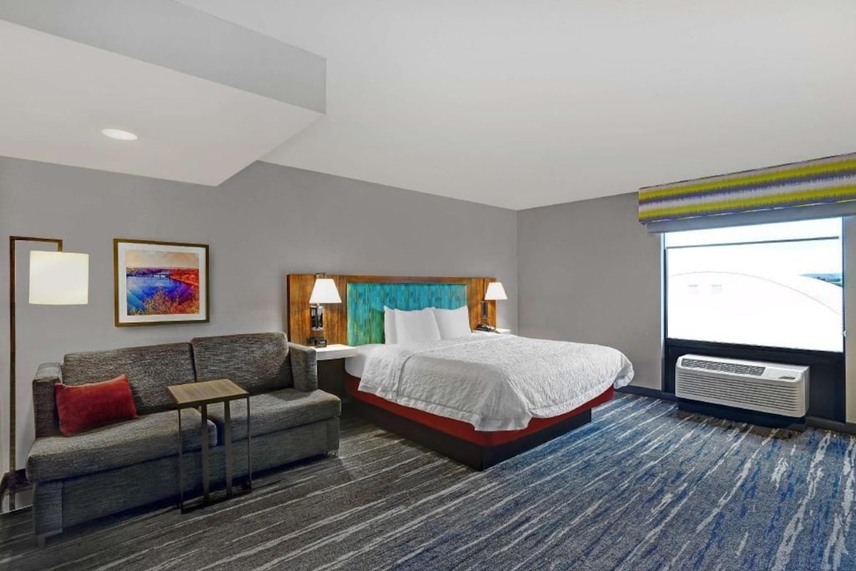 Quarto de hotel com cama, sofá, mesa de cabeceira, luminária, janela com persianas e ar-condicionado fixado na parede.
