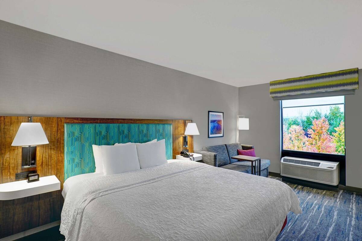 A hotel room with a large bed, two bedside tables with lamps, a small sofa, a window with a view, and an air conditioning unit.
