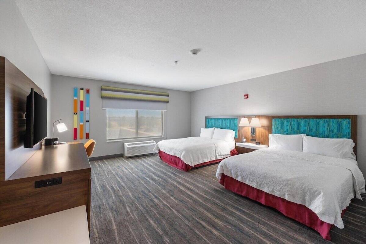 A hotel room with two beds, a desk with a TV, a window, and a wall-mounted air conditioner.