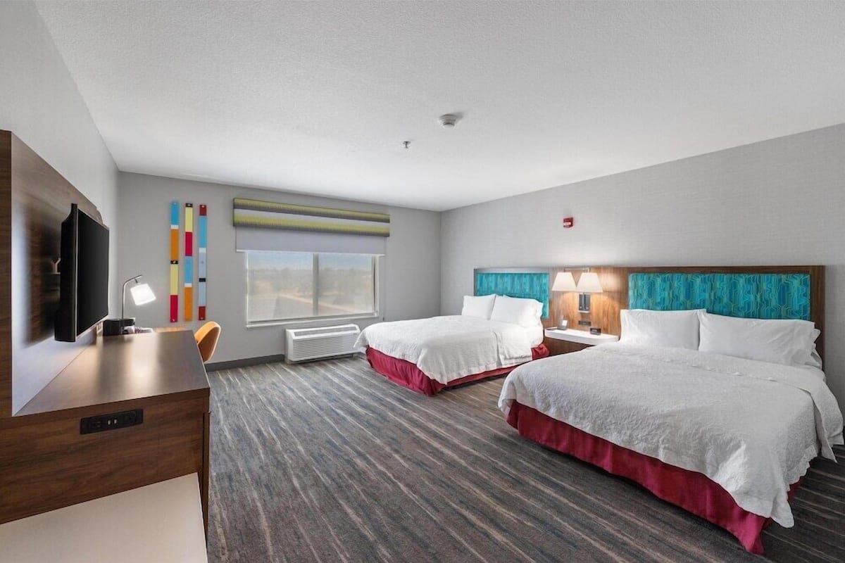 A hotel room with two beds, a desk with a TV, a window, and a wall-mounted air conditioner.