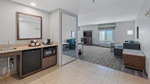 Una habitación de hotel moderna con kitchenette, un espejo grande, una zona de estar con sofá y sillones, y vistas a través de un ventanal.