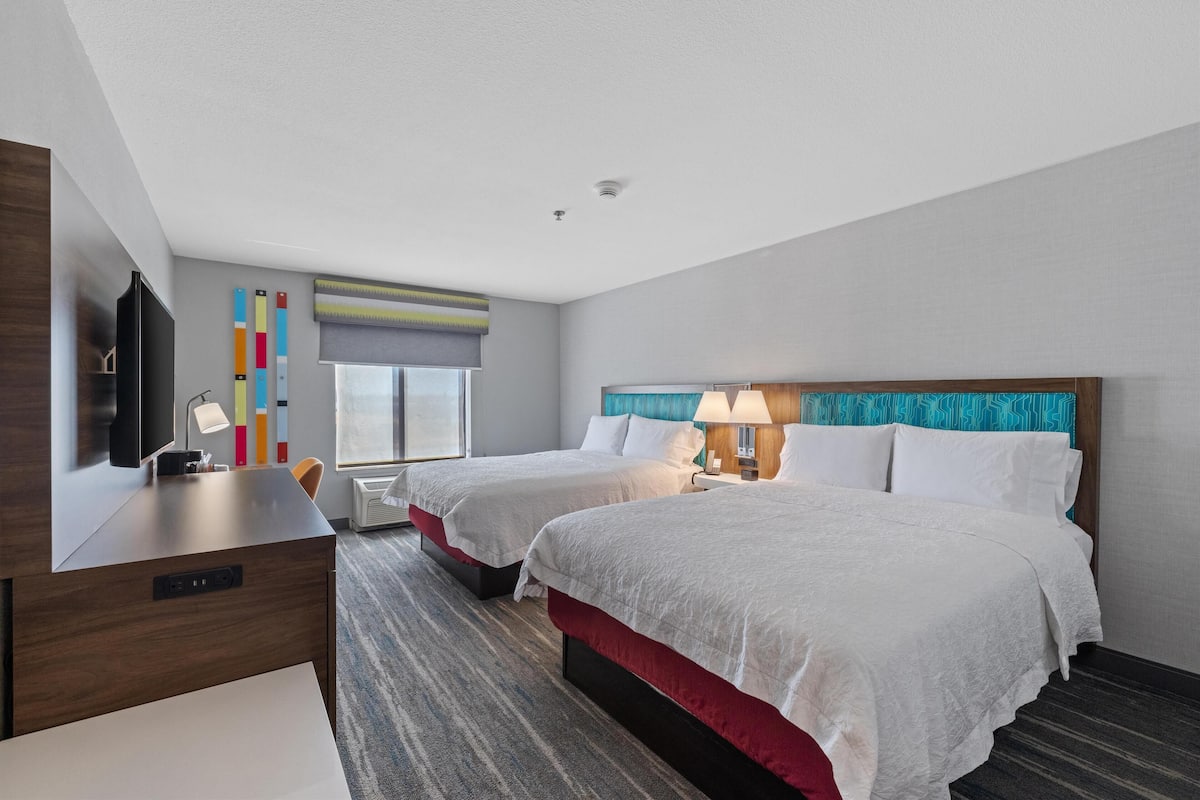 A hotel room with two beds, a desk, a TV, and a window with blinds.