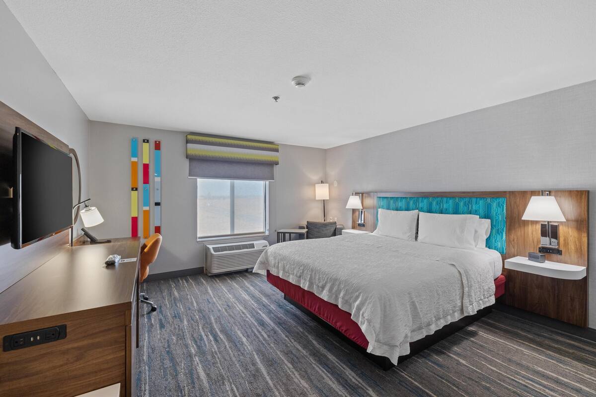 A modern hotel room with a large bed, a desk with a flat-screen TV, a chair, a window with blinds, and a wall-mounted TV.