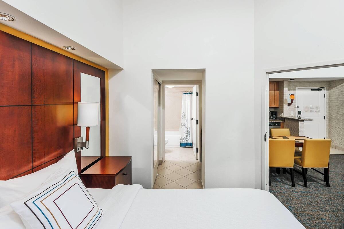 Ein Hotelzimmer mit einem Bett, einem hölzernen Kopfteil, einem Nachttisch und einer Lampe. Eine Tür führt zu einem Badezimmer und einer Küchenzeile mit Tisch und Stühlen.