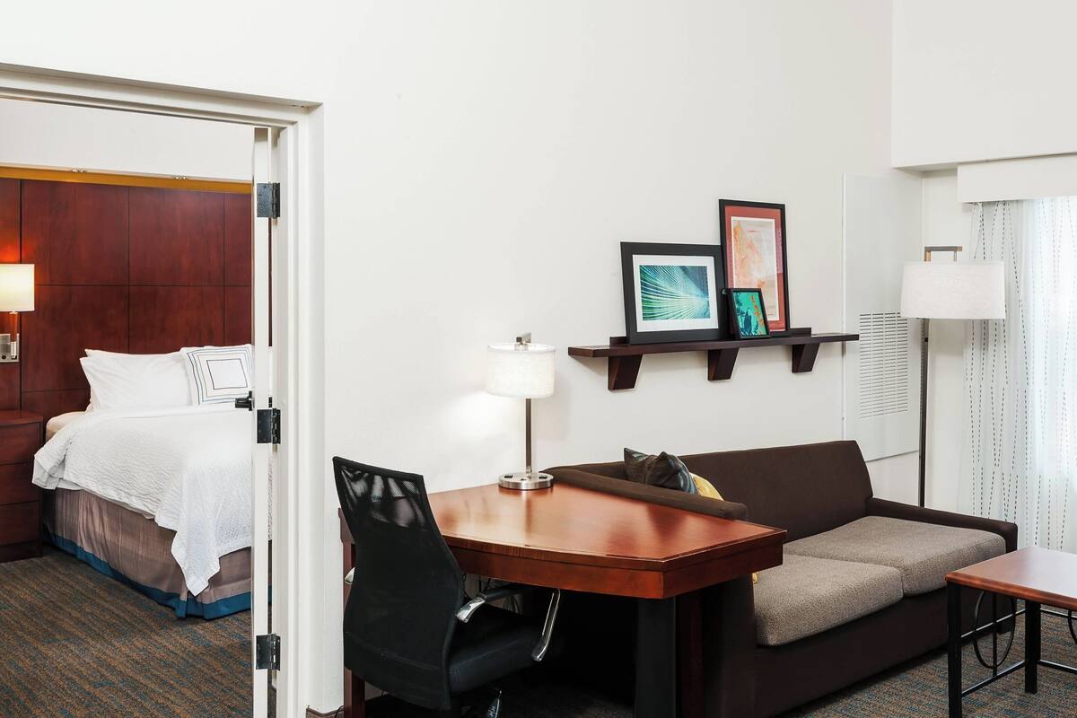 Une chambre d’hôtel avec un lit, un canapé, un bureau et une chaise. Il y a une lampe sur le bureau et un cadre photo au mur.