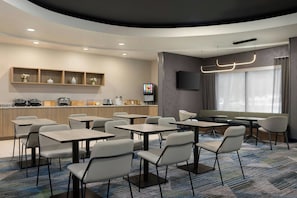 Un comedor moderno de hotel con varias mesas y sillas, una barra de cocina con electrodomésticos y un amplio ventanal con cortinas.