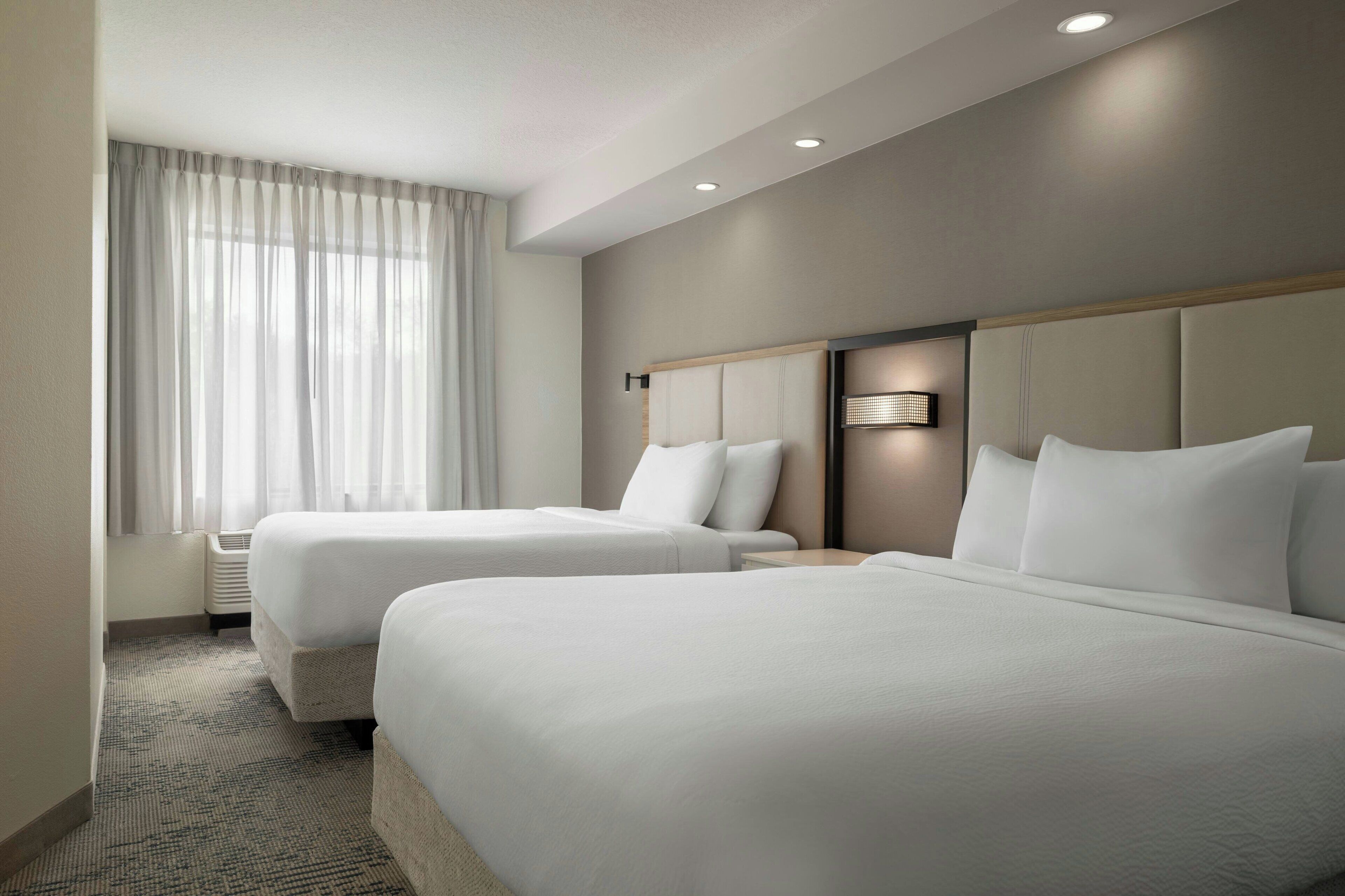 A hotel room with two beds, a large window with curtains, a wall-mounted light, and a carpeted floor.