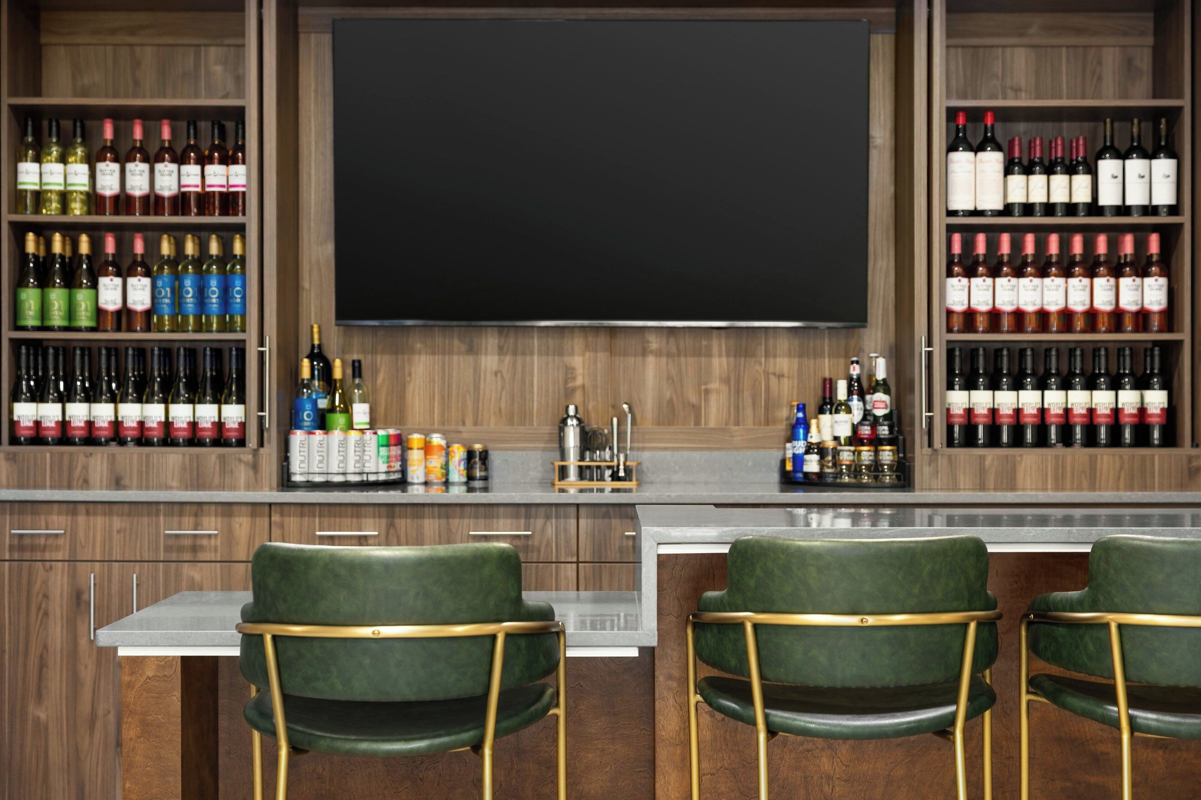 Zona de bar con múltiples estantes llenos de bebidas embotelladas, barra y dos sillones verdes con patas doradas.