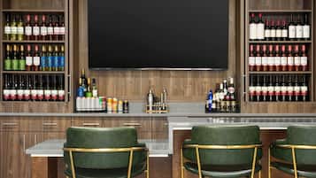 Zona de bar con múltiples estantes llenos de bebidas embotelladas, barra y dos sillones verdes con patas doradas.