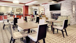 Un comedor amplio con varias mesas y sillas, una zona de bar con distintos electrodomésticos, y una pared de piedra con un televisor montado.