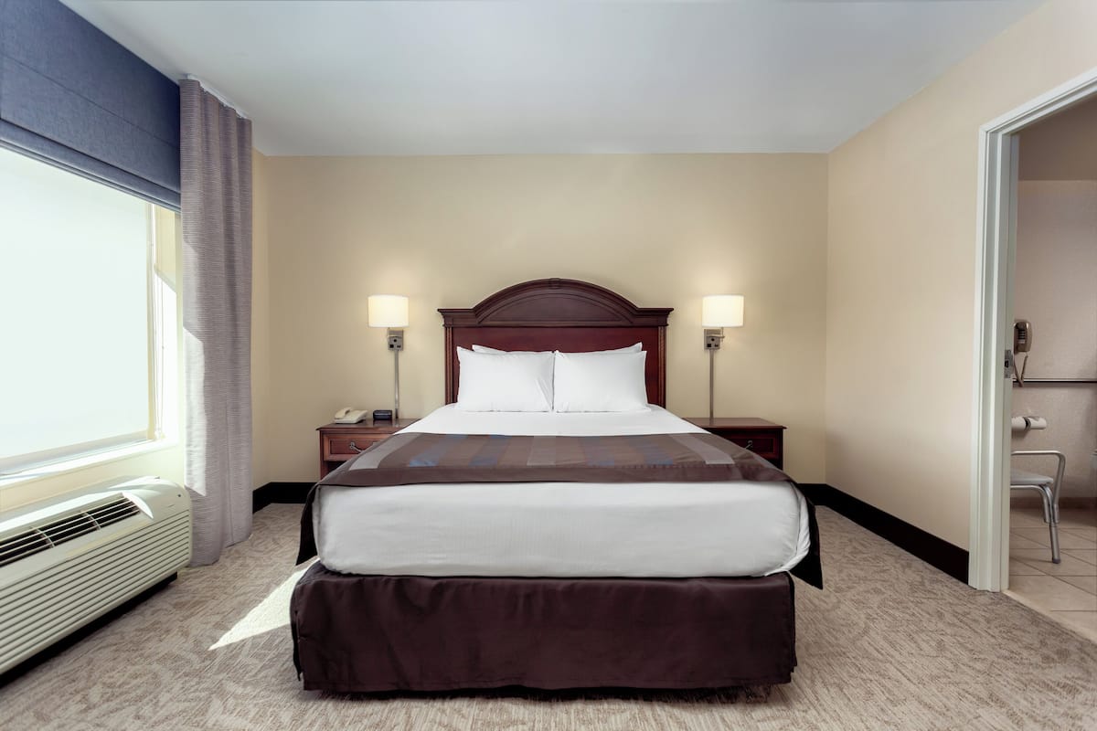 Une chambre d’hôtel avec un grand lit, deux lampes de chevet, une table de nuit, une fenêtre avec des rideaux et une porte donnant sur une salle de bain.