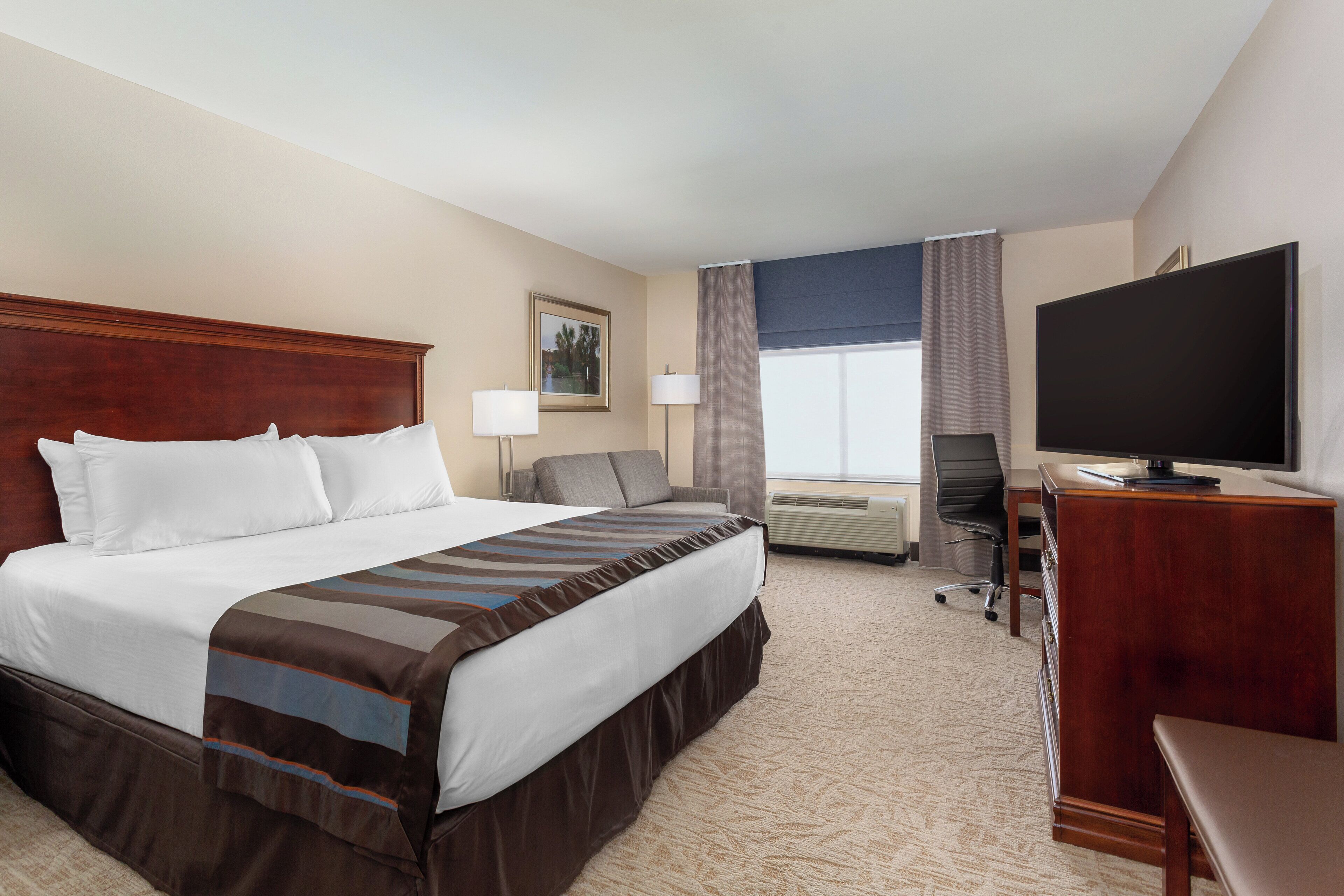 A hotel room with a large bed, a desk with a flat-screen TV, a chair, and a window with curtains.