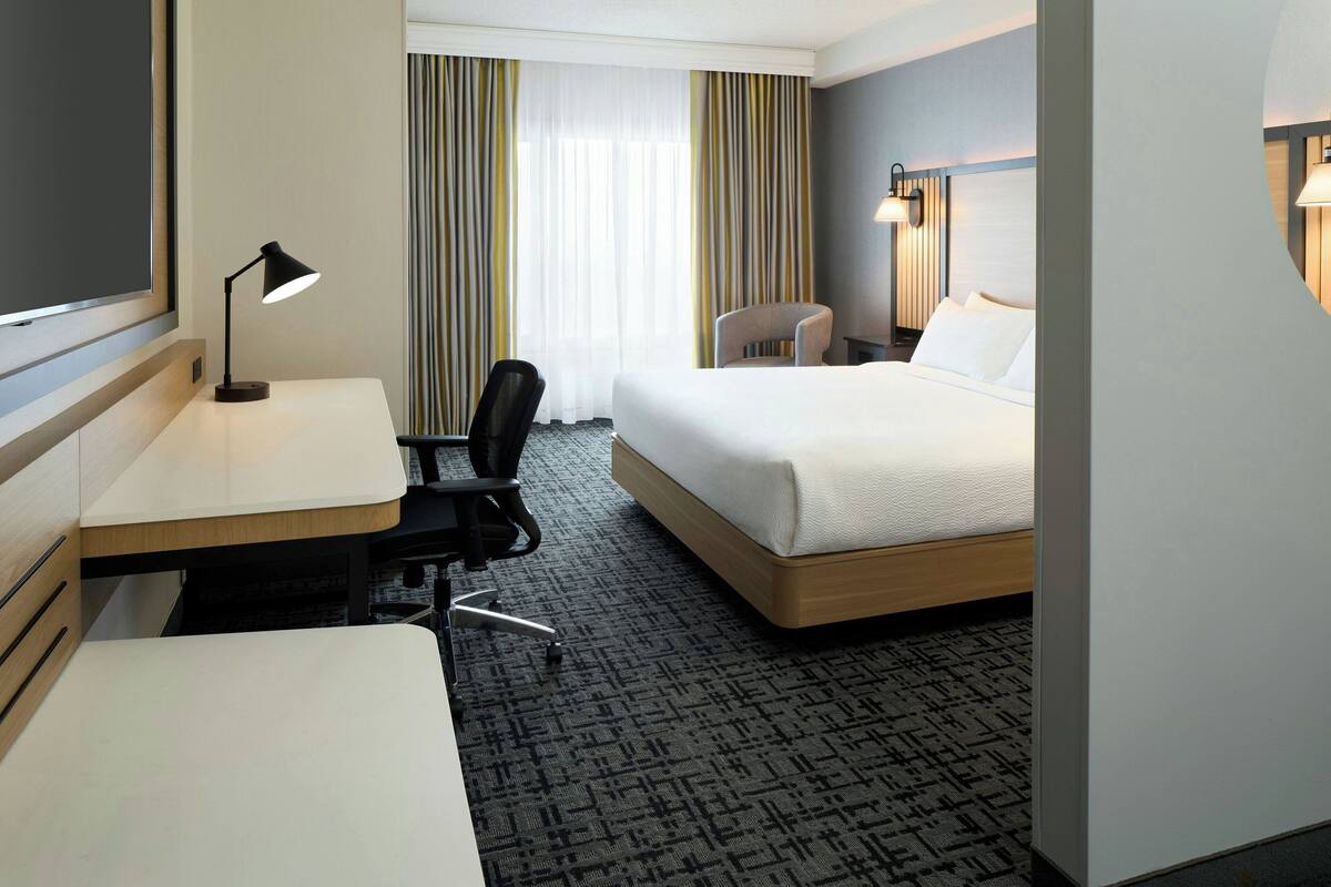Una habitación de hotel con una cama, un escritorio con lámpara, una silla y un gran ventanal con cortinas.