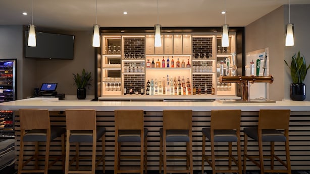 A bar area with a long counter, multiple bar stools, a television mounted on the wall, and shelves stocked with bottles and glasses. - Fairfield Inn & Suites by Marriott Montreal Airport (Dorval)