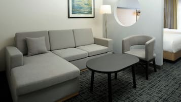 A modern hotel room with a grey sofa, a round table, and a chair. There's a framed picture on the wall and a lamp beside the sofa.