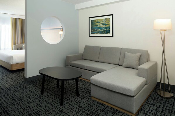 A hotel room with a grey sofa, a round mirror, a small table, a floor lamp, and a painting on the wall.