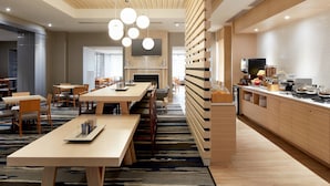 A modern hotel dining area with a long communal table, individual seating, a fireplace, and a kitchen counter with breakfast items. - Fairfield Inn & Suites by Marriott Montreal Airport (Dorval)