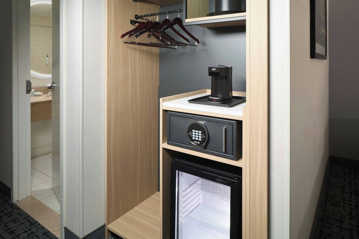 Una habitación de hotel compacta con minibar, cafetera y perchero.