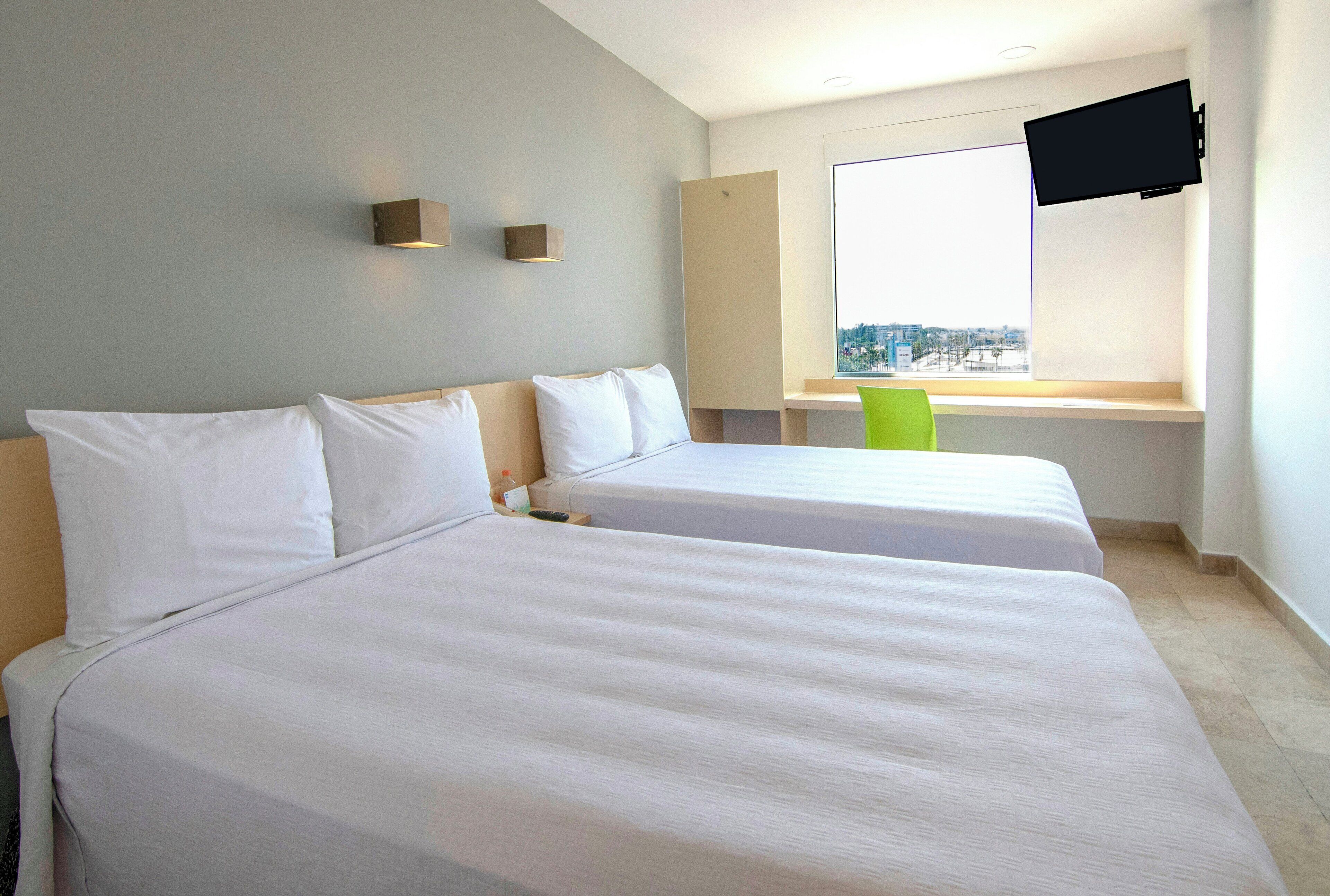 Habitación de hotel con dos camas, un televisor montado en la pared y una ventana con vista a la ciudad.