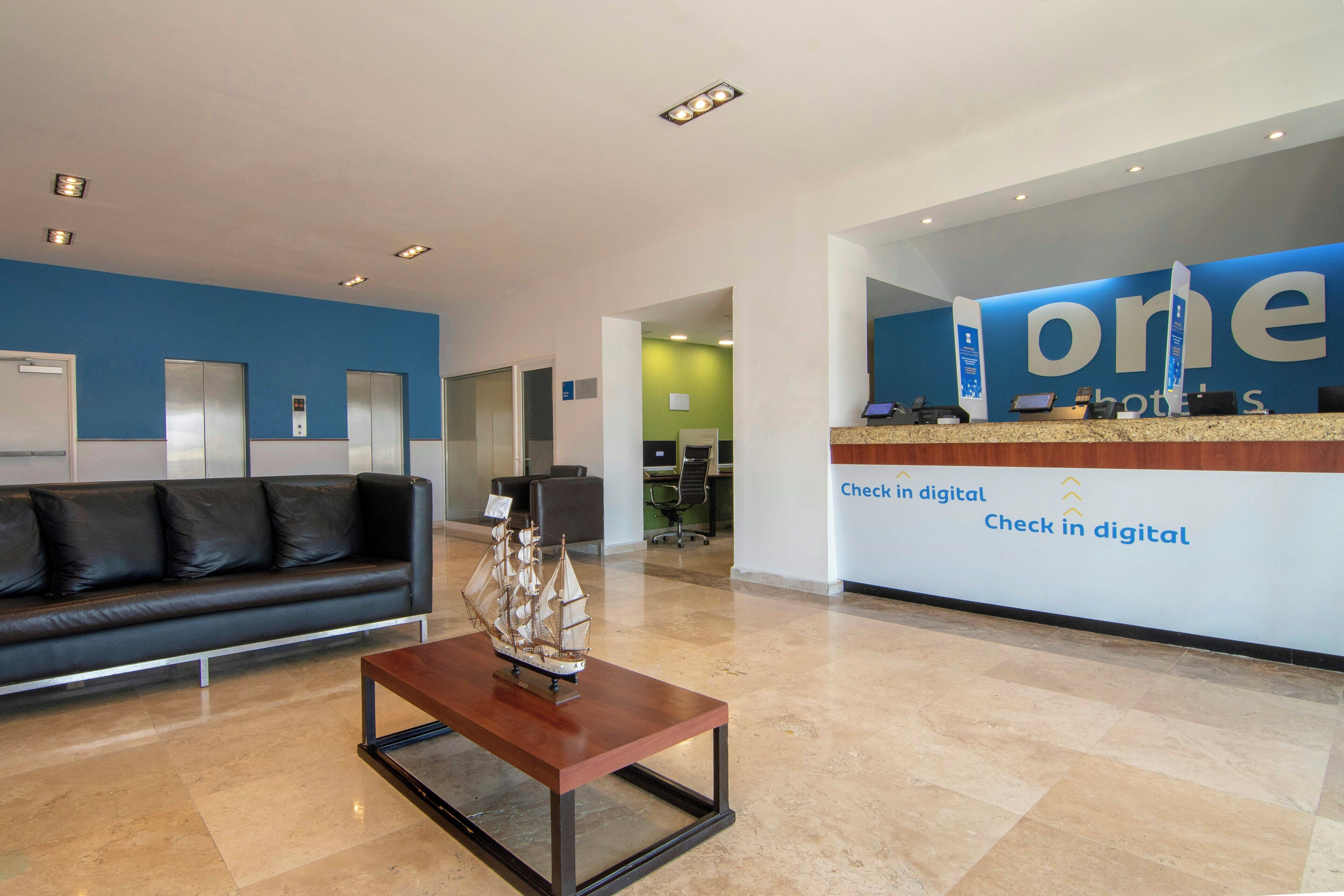Zona de recepción de hotel con un mostrador de registro digital, un sofá de cuero negro y una mesa de centro con un modelo de barco.