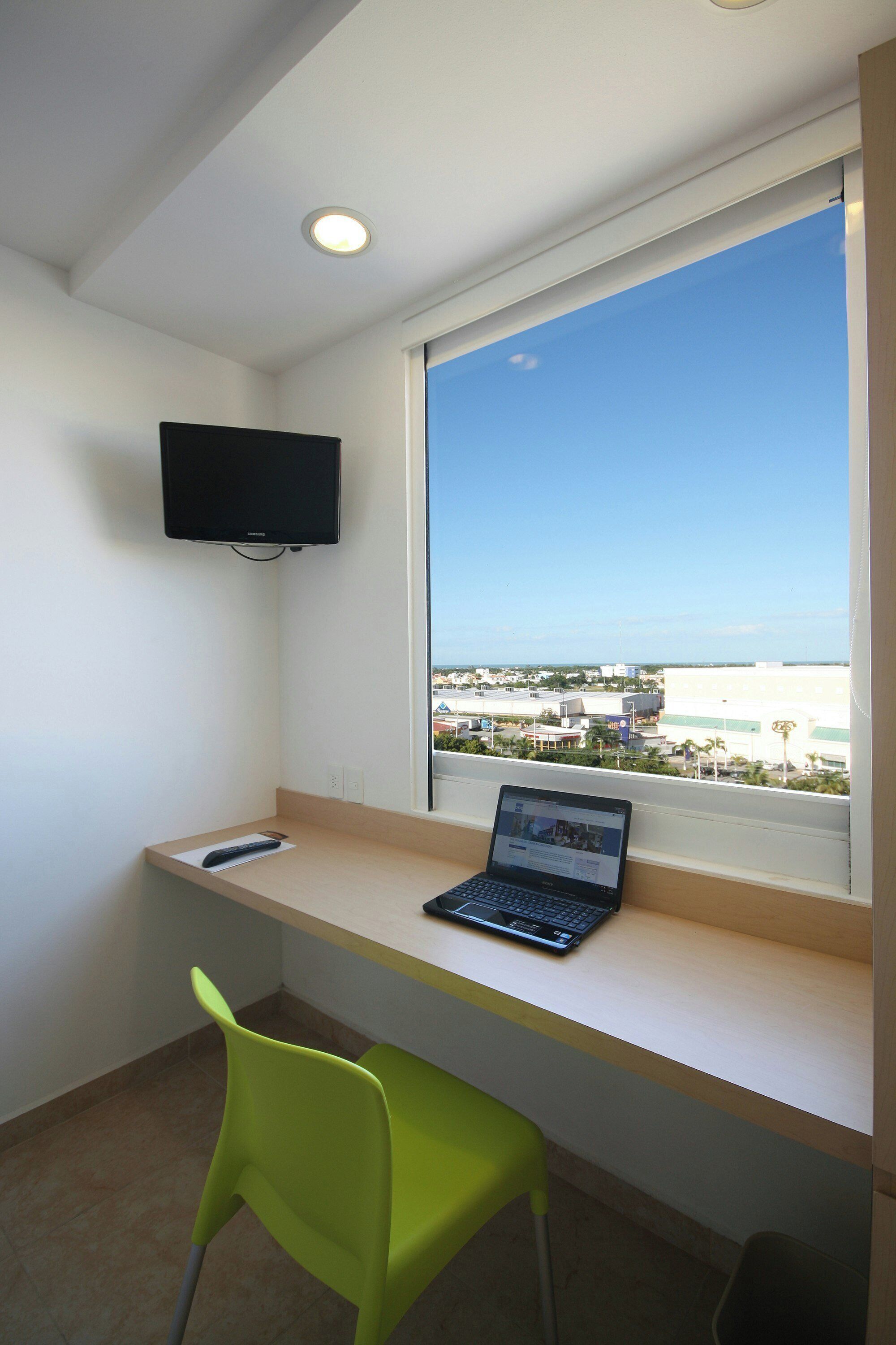 Une pièce comprenant un bureau, un ordinateur portable, une chaise verte, un téléviseur fixé au mur et une fenêtre donnant sur la ville.