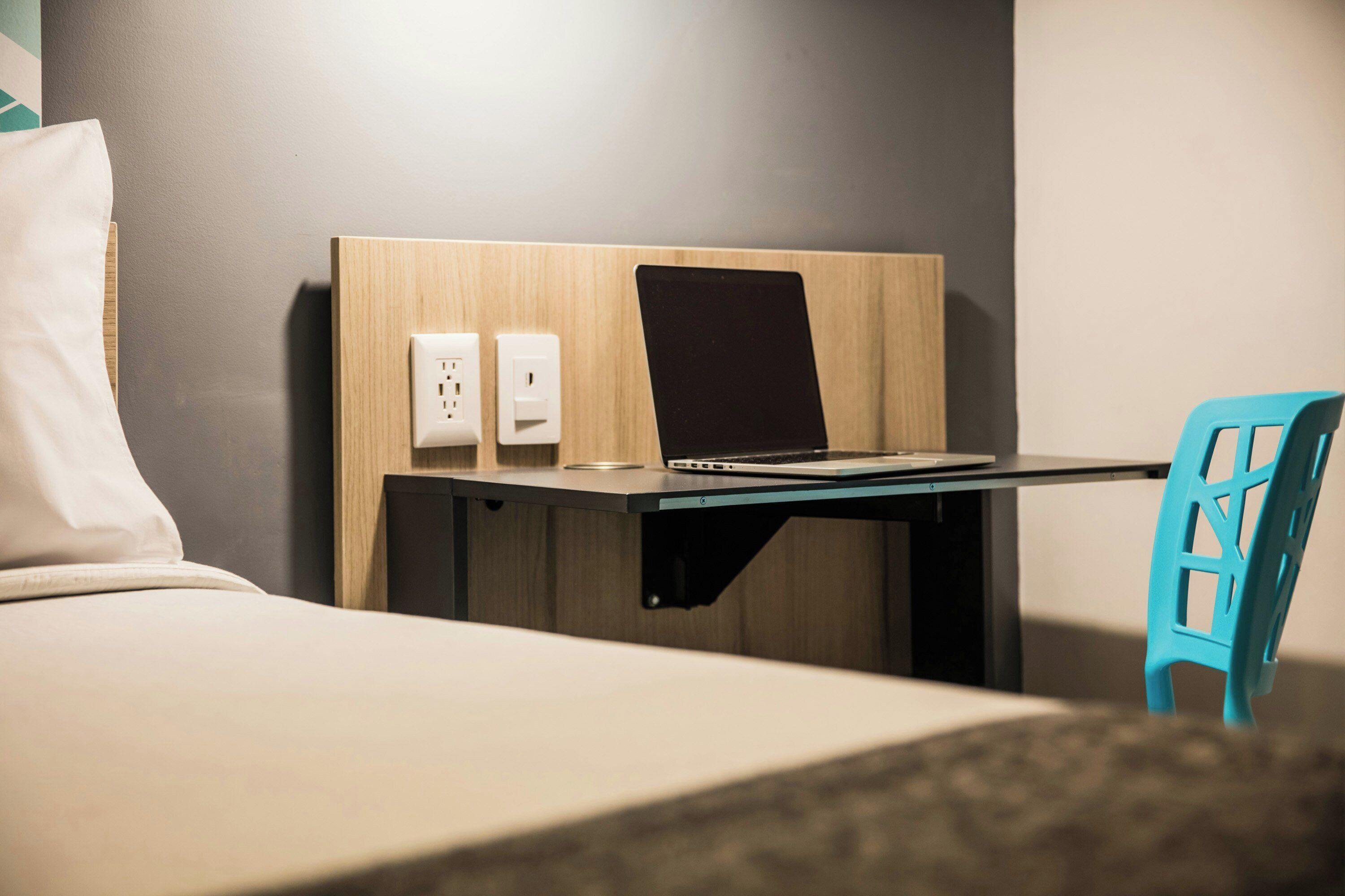 Une chambre d’hôtel avec un lit, un bureau avec un ordinateur portable, une chaise et une prise électrique au mur.