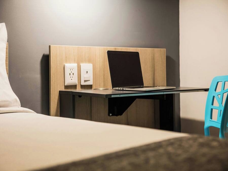 Une chambre d’hôtel avec un lit, un bureau avec un ordinateur portable, une chaise et une prise électrique au mur.