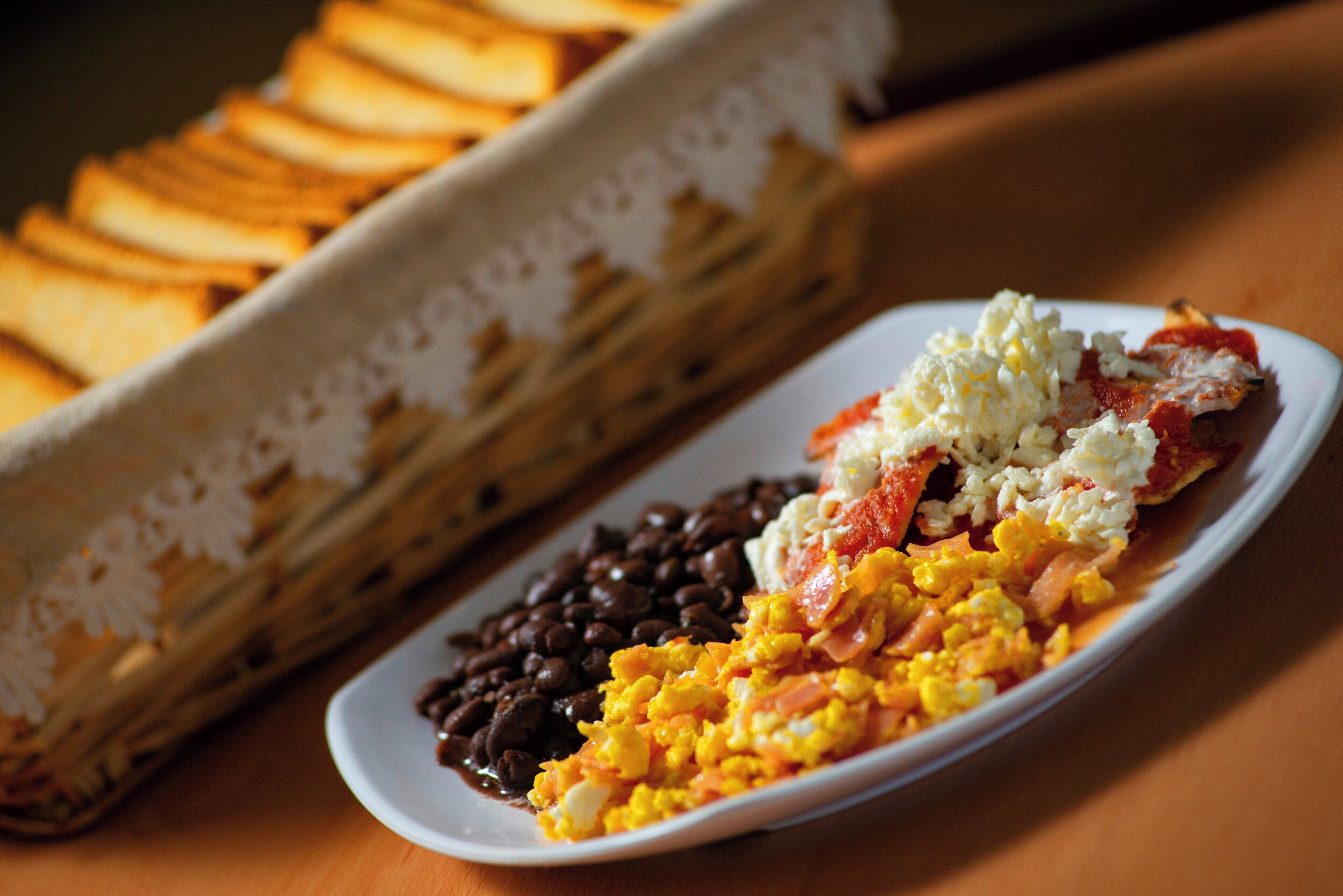 Un plato de comida con frijoles, huevos revueltos y un plato a base de tomate, junto a una bandeja de pan.