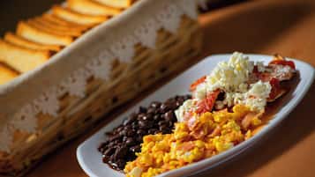 Un plato de comida con frijoles, huevos revueltos y un plato a base de tomate, junto a una bandeja de pan.