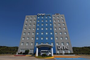 Un hotel con fachada azul y gris, una entrada central con toldo, y una furgoneta estacionada frente a él.