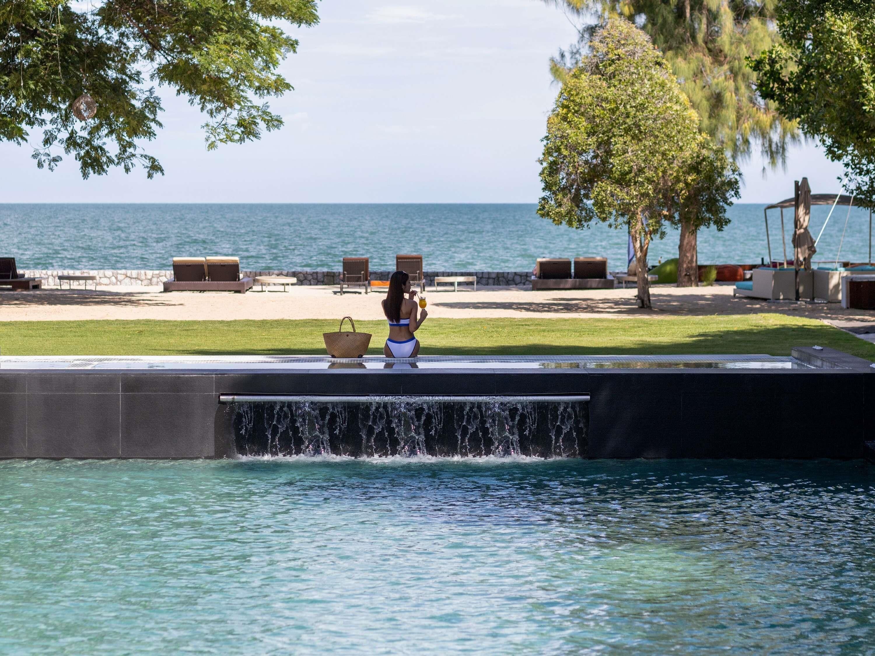 A pool with a water feature, a lounge area with chairs and a table, and a view of the sea.
