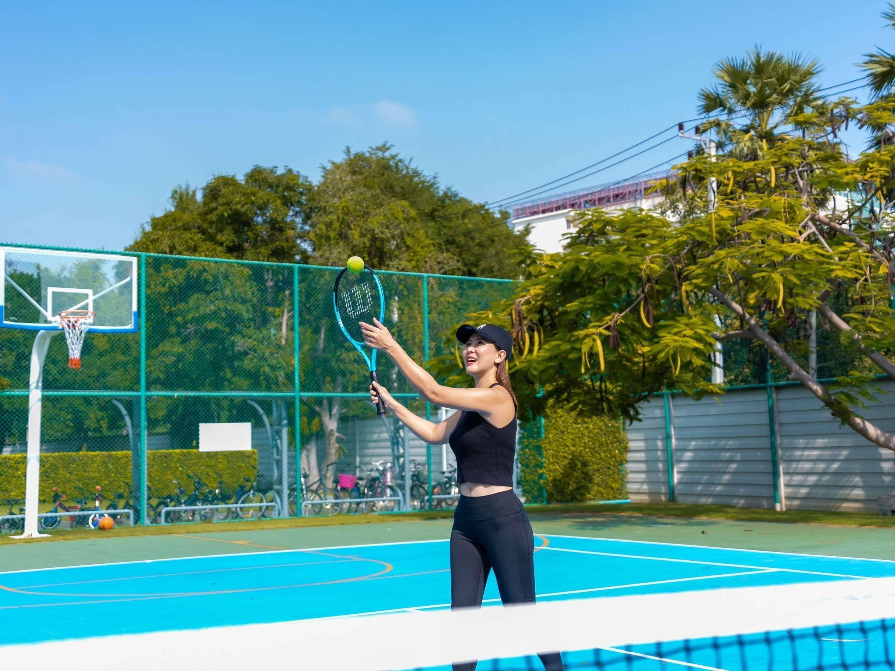 Une personne joue au tennis sur un court extérieur, avec un panier de basketball en arrière-plan.