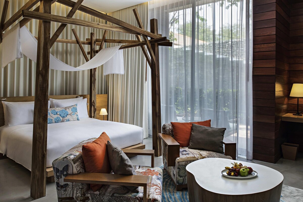 Habitación de hotel con una cama grande, un sofá, una mesa de centro con fruta y vistas al exterior a través de cortinas traslúcidas.