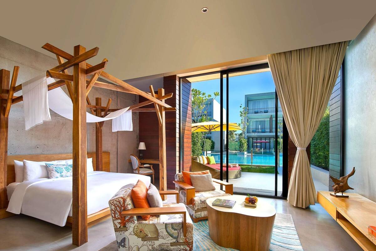 Een moderne hotelkamer met een bed, een houten hemelbed, een zithoek met een ronde tafel en uitzicht op een zwembad en een gebouw.