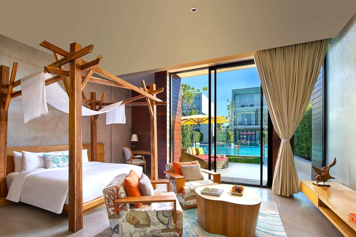 A modern hotel room with a bed, a wooden canopy, a sofa, a round coffee table, and a view of a swimming pool and building.