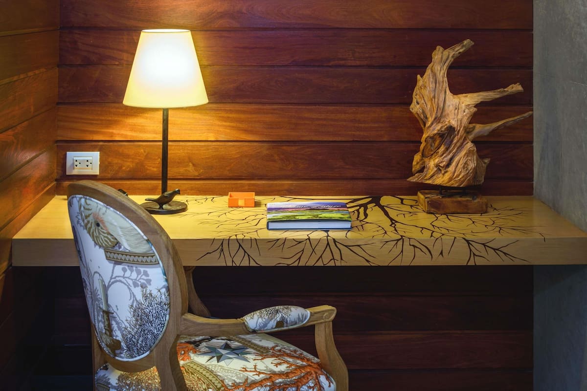 A wooden desk with a lamp, a chair with a patterned cushion, and a decorative tree trunk.