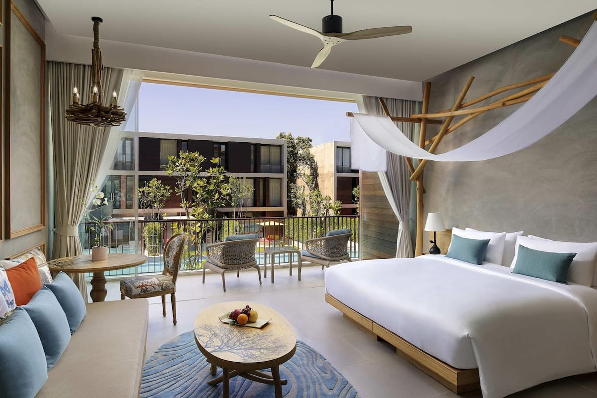 Een hotelkamer met een bed, een ronde tafel met fruit, een bank, een balkon met uitzicht op gebouwen, en een plafondventilator.
